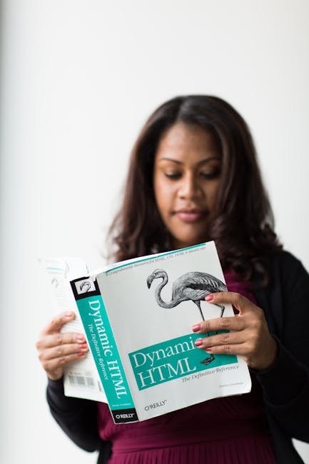 A woman with shoulder-length dark brown hair is holding and reading a book titled 'Dynamic HTML' by O'Reilly, which features a stylized bird illustration on the cover. She is dressed in a dark blazer